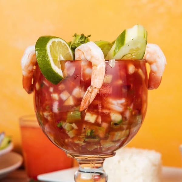 Shrimp cocktail with lime and cucumber in a goblet, with a drink and rice in the background.