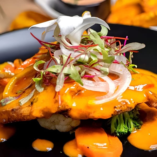 Latin-American dish with orange sauce, microgreens, onions, and a white flower garnish on a black plate.