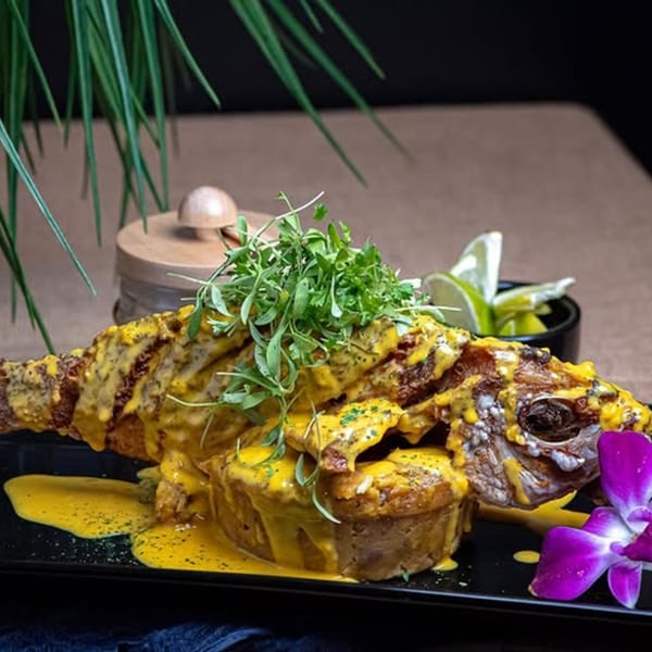 Whole fried fish with yellow sauce and herbs, served with a purple orchid and lime wedges.