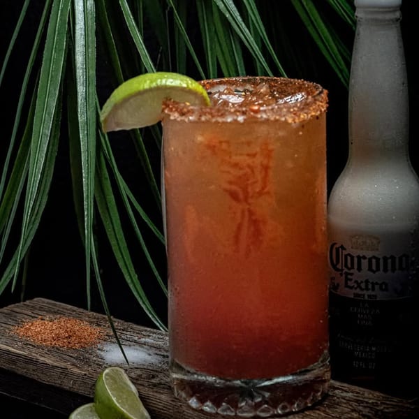 A vibrant red drink with a chili-lime rim and lime wedge, next to a Corona Extra bottle.