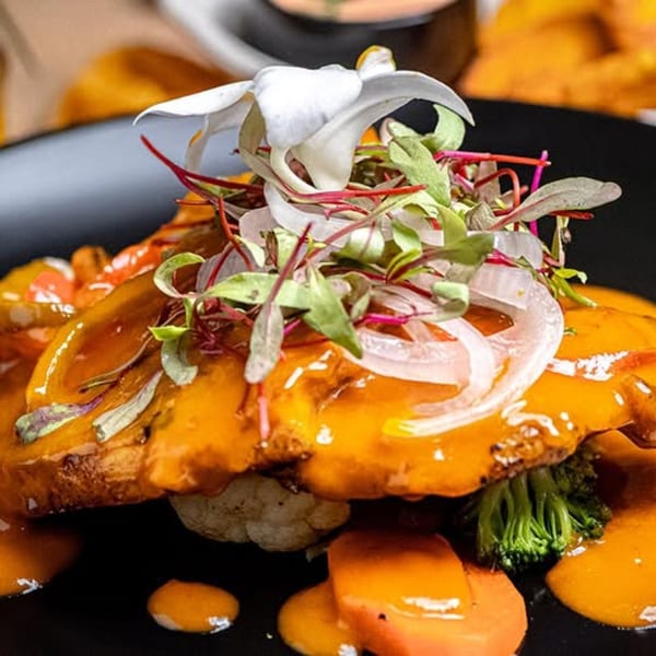 Latin-American dish with orange sauce, microgreens, onions, and a white flower on a black plate.